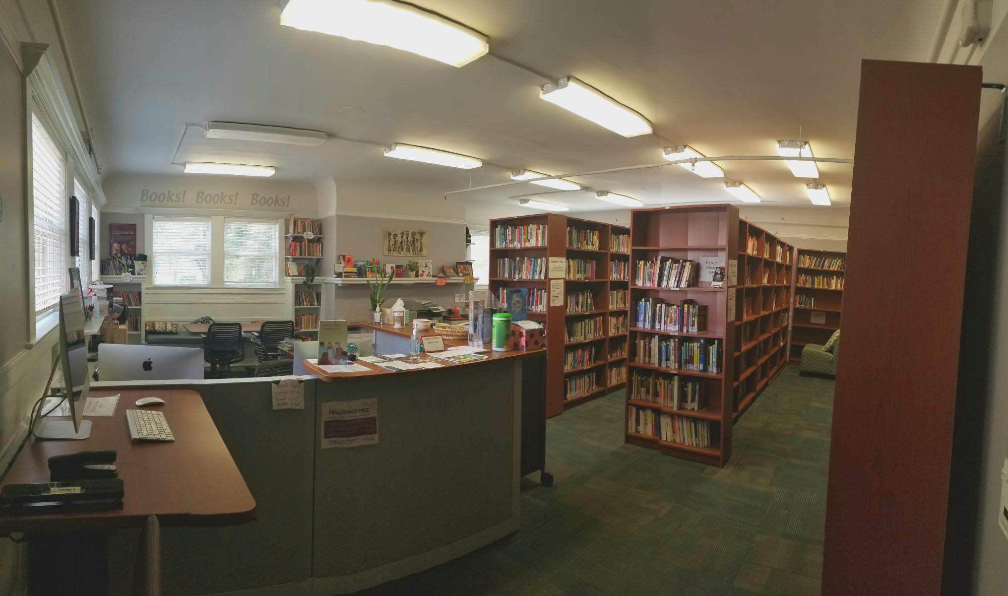 Picture of the inside of the Joy Fergoda library showing the check in desk and rows of books. There are windows in the background and a light olive colored carpet.