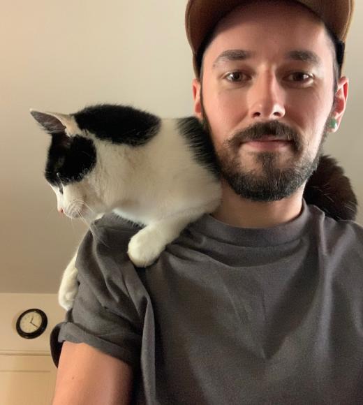 Photo of Aaron with his black and white cat on his shoulder. Aaron has a beard and is wearing a gray t shirt and a baseball hat.
