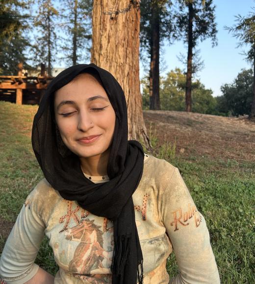 Sara sitting outside smiling with her eyes closed. She is wearing a hijab and a printed shirt.