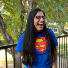 Varuna is wearing a superman shirt and black leggings in front of a fence outside with trees behind the fence. She is wearing glasses with her long hair around her shoulders. Varuna is laughing in the photo with one hand on her leg in front of her and one hand behind her back.